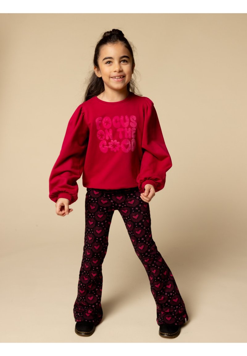 Rode top met lange mouwen en pofmouwen, met de tekst "FOCUS ON THE GOOD". Zwarte uitlopende leggings met een roze hartjespatroon. Zwarte schoenen.