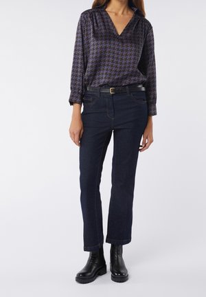 Femme portant un chemisier pied-de-poule noir et bleu, un jean bleu foncé avec ceinture noire, et des bottines noires, sur un fond uni.