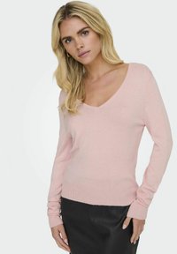 Hellrosa V-Ausschnitt-Pullover aus weichem Stoff, mit langen Ärmeln und gerippten Bündchen. Tragen mit einem schwarzen Lederrock.