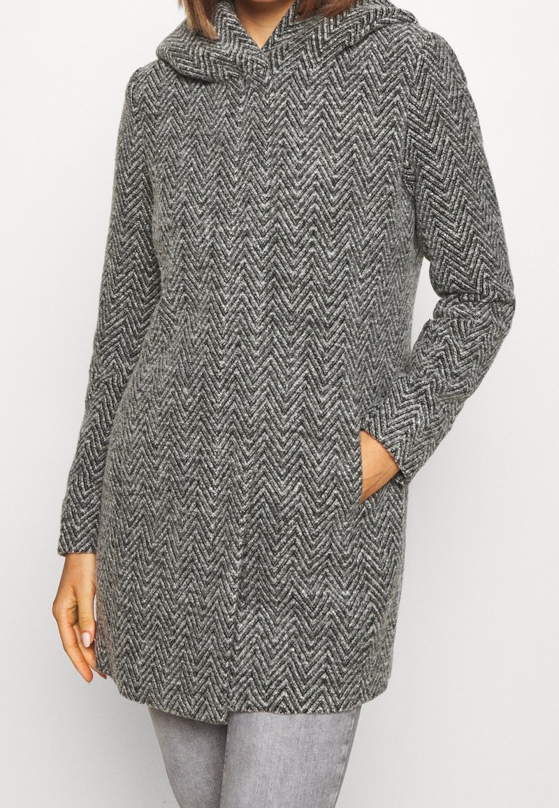 Femme portant un manteau en laine gris à motif chevrons avec un col montant et des poches latérales, associé à un pantalon gris clair.