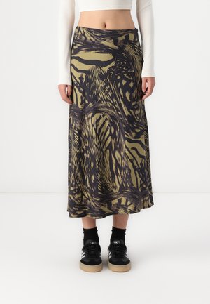 Jupe longue en imprimé animal vert et noir, fabriquée en tissu lisse, avec un design fluide et un ourlet irrégulier, associée à des baskets noires.