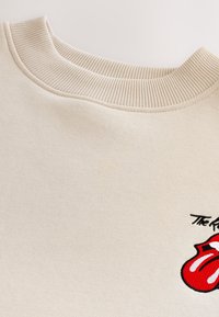 Krämfärgad sweatshirt med ribbad krage och mjuk textur. Har en röd och svart Rolling Stones-logga på framsidan.