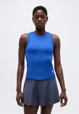 Atletisk kvinde iført en ærmeløs blå top og mørkegrå tennisnederdel, stående med armene afslappet mod en ensfarvet lys baggrund.