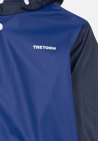 Granatowa i ciemnoniebieska kurtka przeciwdeszczowa o gładkiej fakturze. Posiada logo "TRETORN" w białym kolorze na froncie oraz białe zatrzaski.
