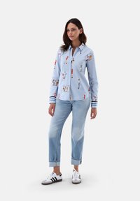 Blue button-up shirt featuring colorful printed figures, long sleeves with striped cuffs, paired with light blue rolled-up jeans and sneakers.