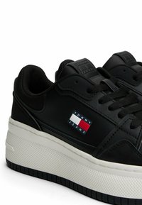 Sneaker in pelle nera con suola in gomma bianca, dotate di un etichetta logo a colori rosso, bianco e blu sul lato.