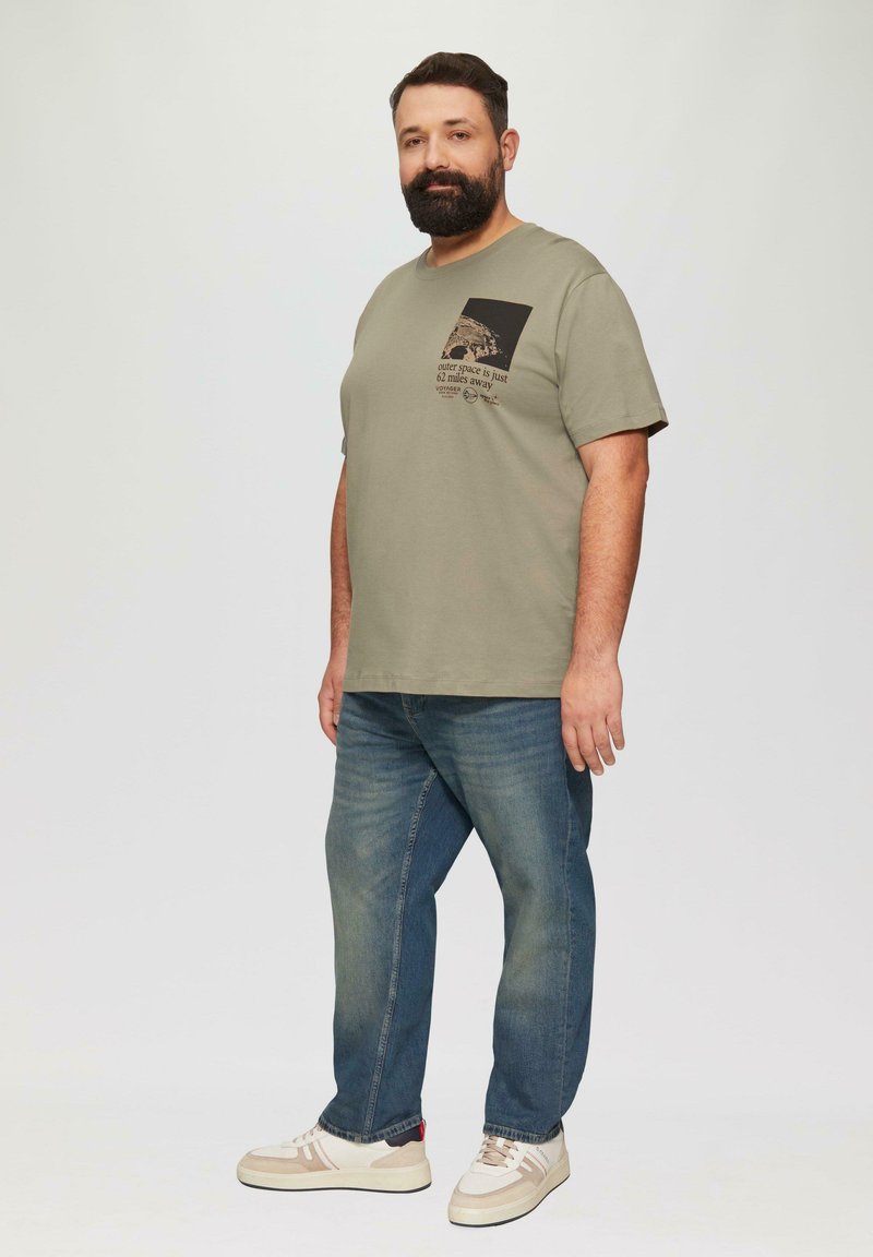Olijfgroen T-shirt met korte mouwen en een zwarte vierkante zak met grafische tekst. Draagt met blauwe spijkerbroek en crèmekleurige sneakers.
