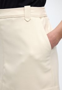 Κοντινή λήψη ενός χεριού που ακουμπά στην μπροστινή τσέπη μιας κρεμ φούστας με θηλιές για ζώνη.