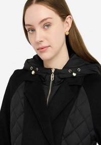 Cappotto nero con cappuccio trapuntato, chiusura con zip dorata e dettagli con ganci. Presenta una combinazione di materiali lisci e testurizzati.