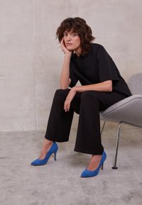 Svart outfit med korta ärmar och vida byxor, matchat med blå mockastövlar med hög klack. Individ som sitter på en modern grå stol.