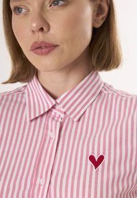 Chemise rayée rose et blanche avec un col boutonné, comportant une petite broderie en forme de cœur rouge sur le côté gauche. Matériau en coton et texture lisse.