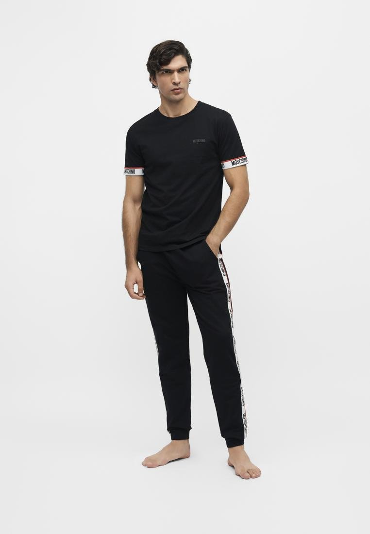 Homme debout pieds nus portant un t-shirt noir Moschino et un jogging assorti avec le logo de la marque sur les manches et sur les côtés du pantalon.