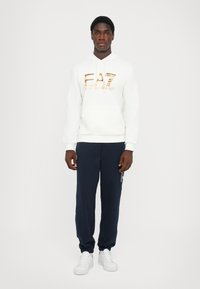 Sweatshirt branca com o logótipo dourado "EA7", combinada com calças de fato em azul-marinho. As calças têm uma marcação lateral branca. O modelo usa ténis brancos.