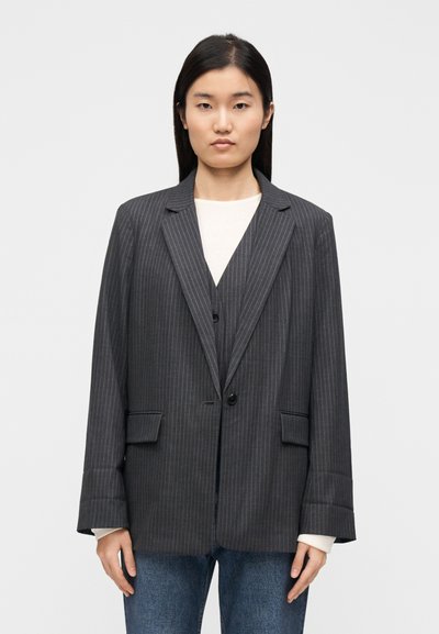 Blazer gris à fines rayures à coupe décontractée, doté de revers crantés, d'une fermeture à un bouton et de deux poches frontales. Porté sur un haut blanc.