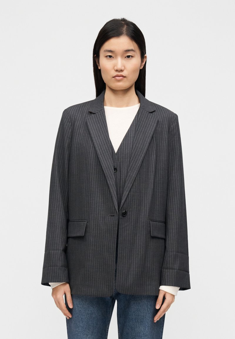 Blazer gris à fines rayures à coupe décontractée, doté de revers crantés, d'une fermeture à un bouton et de deux poches frontales. Porté sur un haut blanc.