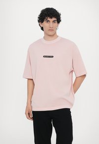 T-shirt de algodão rosa claro com mangas curtas, decote redondo e um patch retangular preto com o logotipo "MICHAEL KORS" na frente.