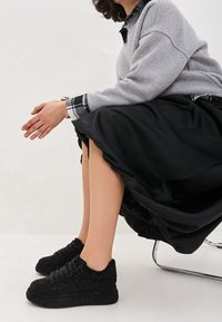 Czarne teksturowane sneakersy z błyszczącym wykończeniem, z okrągłym noskiem i sznurowaną konstrukcją, noszone z czarną satynową spódnicą i szarym swetrem.