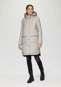 Grijze pufferjas met een fleece onderstuk, voorzien van een capuchon, een knopenfront en twee voorzakken. Draag het over zwarte broek en laarzen.