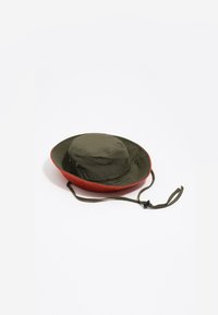 Odwracalna czapka typu bucket w oliwkowym kolorze z górną częścią oraz czerwonym, teksturowanym wnętrzem, otworami wentylacyjnymi oraz regulowanym strapem na brodzie z zapięciem typu toggle.