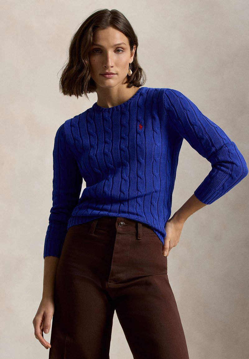 Blå kabelstrikket genser med rund nakke og trekvart ermer, med en liten rød logo. Kombinert med brune bukser med høyt liv.