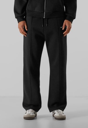 Homme portant un pantalon de survêtement noir à coupe décontractée et des baskets blanches, debout devant un fond uni.