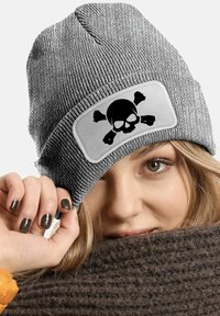 Gray knitted beanie with a large, white rectangular patch featuring a black skull and crossbones design. Textured fabric and fitted shape.