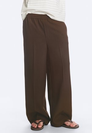 Personne portant un pantalon marron ample avec une taille élastique et des tongs foncées, debout sur un fond clair uni.