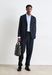 Navy tailored suit jacket and pants, light blue shirt, black shoes, holding a gray structured bag with top handles. Smooth fabric, minimalist design.