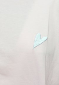 Tessuto di cotone rosa chiaro con una morbida texture, caratterizzato da un piccolo dettaglio di cuore blu ricamato sul lato sinistro.