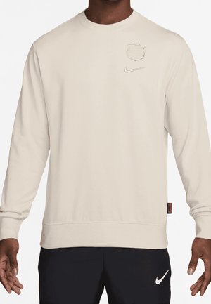Mann trägt beige Sweatshirt mit langen Ärmeln, schwachem Logo und Nike-Swoosh auf der Brust, kombiniert mit schwarzen Nike-Hosen mit weißem Swoosh.