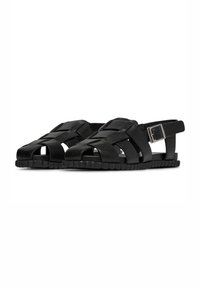 Sandalia negra con diseño de cuero trenzado, punta abierta, correa ajustable en el tobillo y suela texturizada. Presenta un acento de hebilla rectangular plateada.