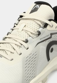 Chaussure de sport de couleur claire avec une tige en mesh respirant, des lacets plats et un logo noir distinctif. Accent Deltastrap remarquable.