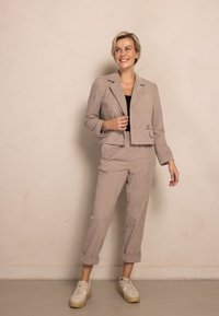 Gekürzte beige Cord-Blazer mit Ein-Knopf-Verschluss und aufgesetzten Taschen, kombiniert mit passenden umgeschlagenen Hosen und weißen Plateausneakern.