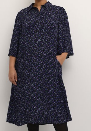 Femme portant une robe fleurie sombre jusqu'aux genoux avec des manches trois-quarts, un col et des poches, debout contre un mur clair uni.