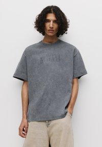 Grå oversized t-shirt med en vasket tekstur, prydet med et subtilt præg af "NIRVANA" logoet på brystet og en rund halsudskæring.