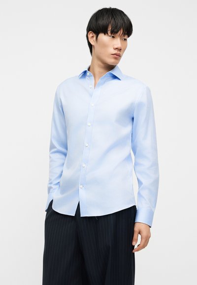 Νέος άνδρας που φοράει ανοιχτό μπλε πουκάμισο με κουμπιά και μακριά μανίκια, καθώς και σκούρο παντελόνι με λεπτές ριγέ γραμμές, κοιτάζει στο πλάι με φόντο ένα απλό φόντο.