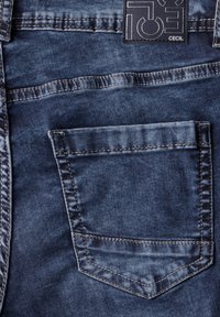 Jean en denim avec un délavage bleu foncé, comportant une poche arrière et des coutures contrastées. Étiquette de marque "CECI" visible au niveau de la taille.