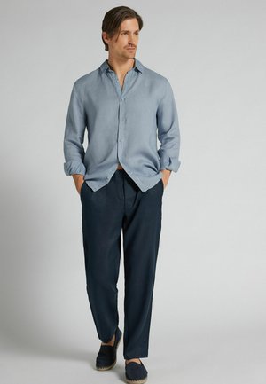 Mężczyzna stoi z rękami w kieszeniach, ubrany w jasnoniebieską koszulę zapinaną na guziki, ciemnogranatowe spodnie i granatowe wsuwane buty, na jednolitym tle.