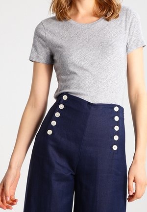 Kobieta ubrana w szarą koszulkę z krótkim rękawem włożoną do granatowych spodni z wysokim stanem z dwoma pionowymi rzędami białych guzików.