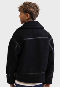 Schwarze Shearlingjacke mit kragenförmigem Ausschnitt, strukturiertem Stoff, Lederelementen und einer Schnallendetail in der Taille.