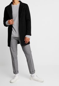 Schwarzer Wollmantel, grauer Sweatshirt und graue karierte Hose. Weiße Turnschuhe. Lange Ärmel, aufgekrempelte Manschetten und sichtbare Knöpfe am Mantel.