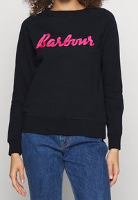 Sweat-shirt noir avec logo "Barbour" en cursive rose, fabriqué dans un tissu doux, doté de poignets et d'un ourlet côtelés, avec une coupe décontractée. Un jean bleu est visible en dessous.