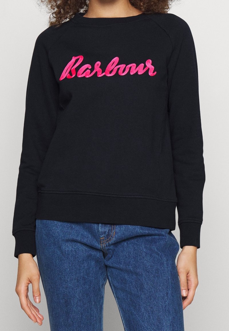 Sweat-shirt noir avec logo "Barbour" en cursive rose, fabriqué dans un tissu doux, doté de poignets et d'un ourlet côtelés, avec une coupe décontractée. Un jean bleu est visible en dessous.