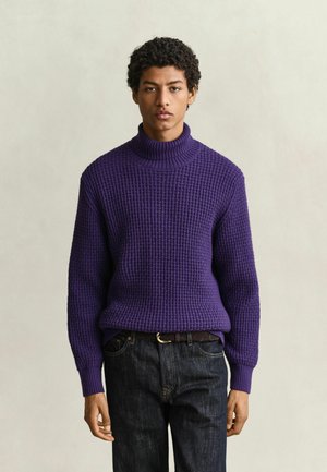Giovane uomo che indossa un maglione a collo alto viola strutturato e jeans scuri, in piedi davanti a uno sfondo chiaro e uniforme.