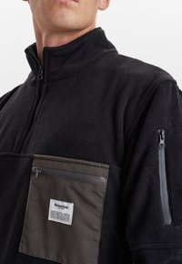 Sort fleece-trøje med høj krave, har en grå brystlomme med lynlås og mærkelogo, samt en lomme med lynlås på ærmet.