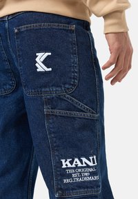 Denim cargobyxor i mörkblått, med en framträdande vit broderad logotyp och ytterligare fickor. Hållbart material och strukturerad design.