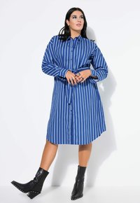 Robes chemises à rayures bleues avec fermeture à boutons et ceinture, manches longues, atteignant le milieu de la cuisse. Portée avec des bottines noires à la cheville en cuir texturé.