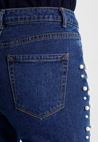 Denimjeans i mörkblått med en strukturerad yta. Har en bakficka och vita pärlprydnader längs sid sömmen.
