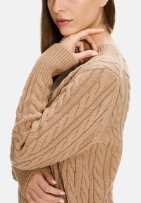 Beige Kabelstrickpullover mit strukturiertem Muster, langen Ärmeln und geripptem Kragen. Weicher Stoff mit lässiger Passform, ausgestattet mit breiten Bündchen.