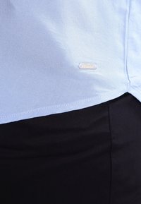 Ljusblå skjorta av vävd tyg, med en liten metallskylt, fållade kanter och en mjuk textur mot svarta byxor.